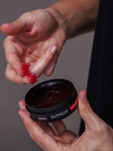 “Tangan mengambil pomade water-based dari jar dengan gelas air dan sisir di latar”